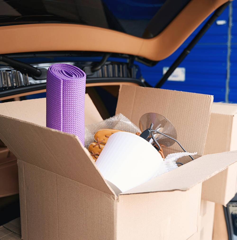 An open cardboard box filled with household items, including a purple yoga mat, a white lampshade, and a disassembled light fixture, placed in the trunk of a car with a blue storage unit in the background.