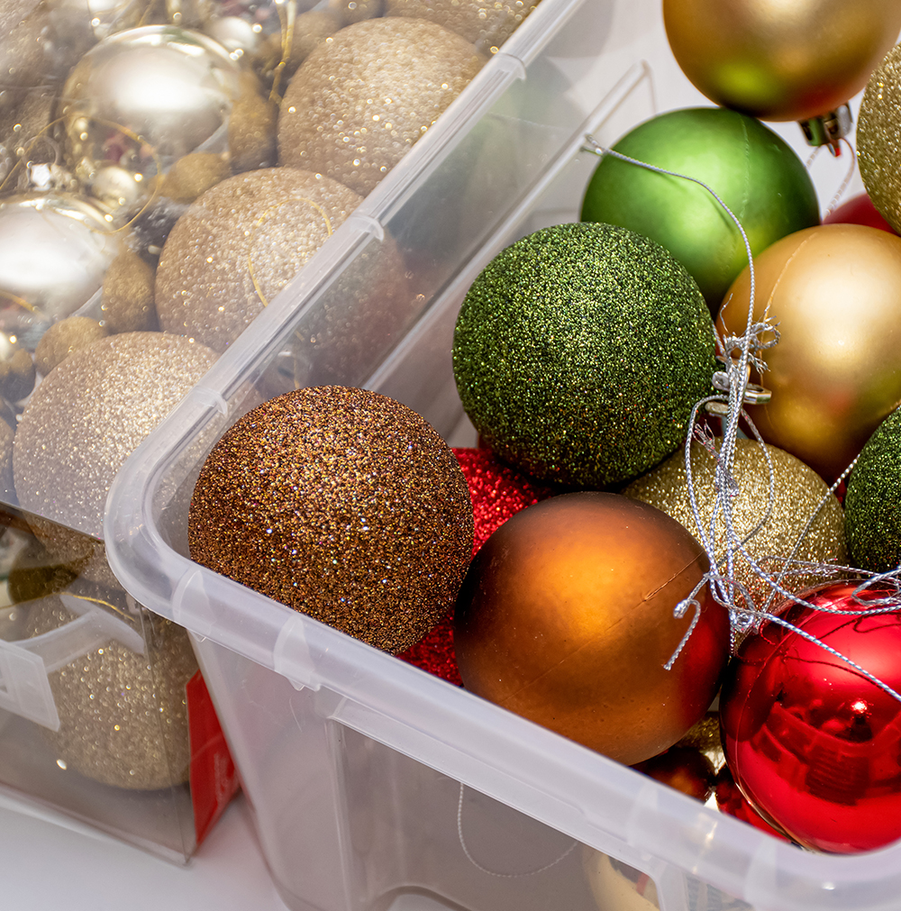 A close-up view of transparent plastic storage boxes filled with Christmas ornaments in various colors, including gold, green, red, and bronze.