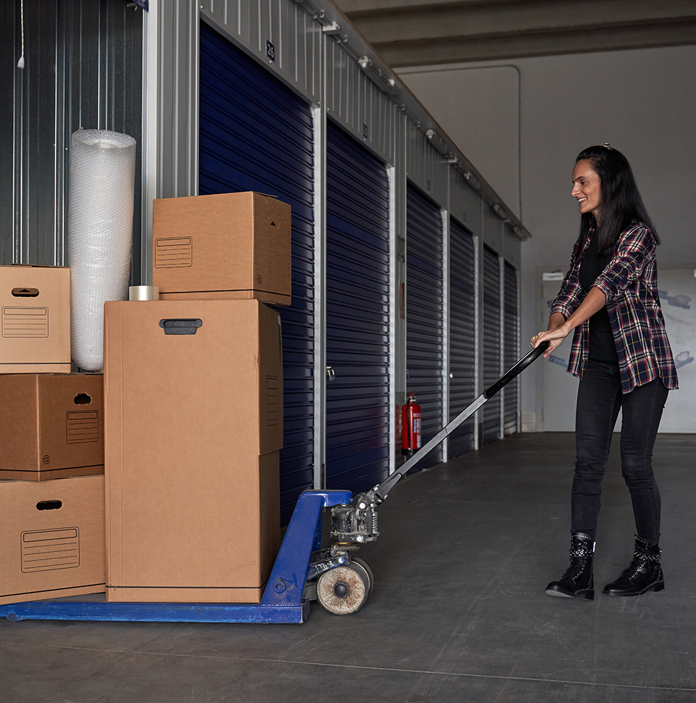 A smiling woman in a plaid shirt and black jeans uses a pallet jack to move stacked cardboard boxes in a storage facility with blue roll-up doors.