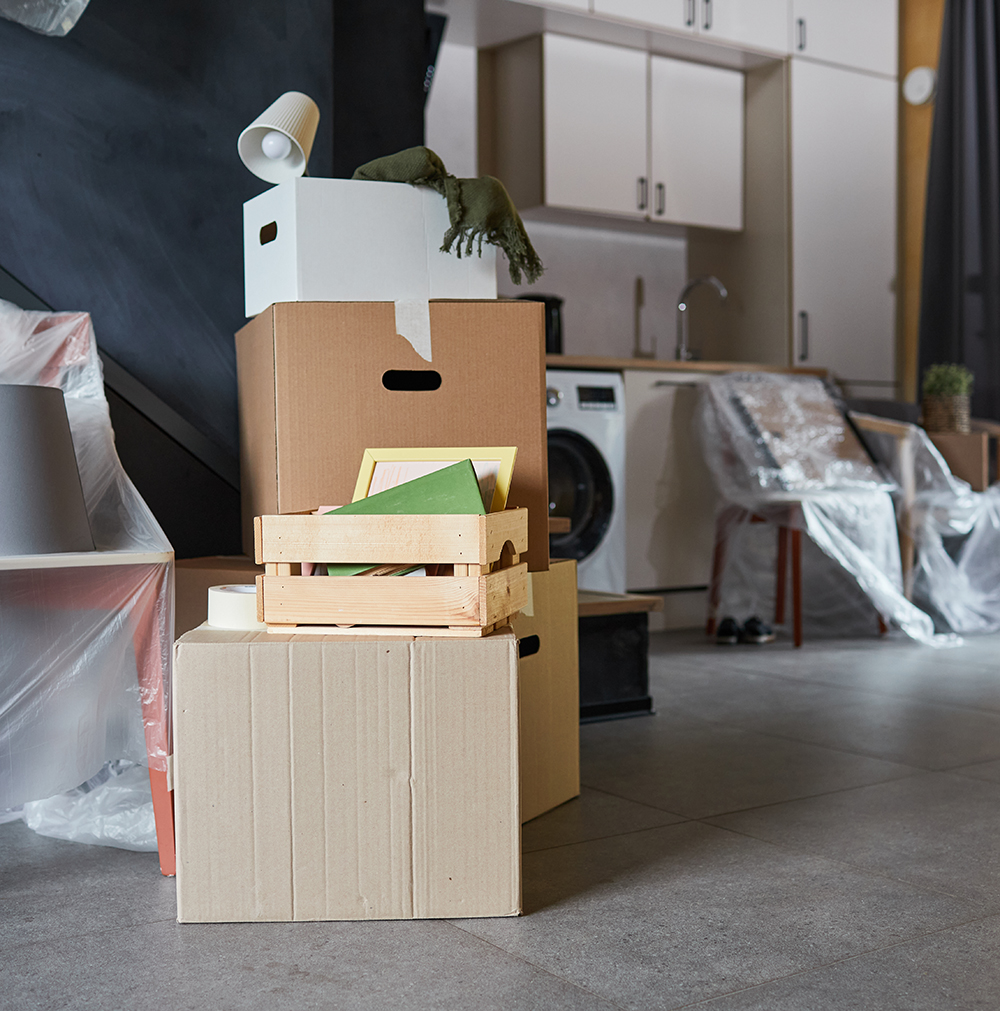 A stack of moving boxes in the middle of a modern kitchen, with some furniture covered in plastic in the background.