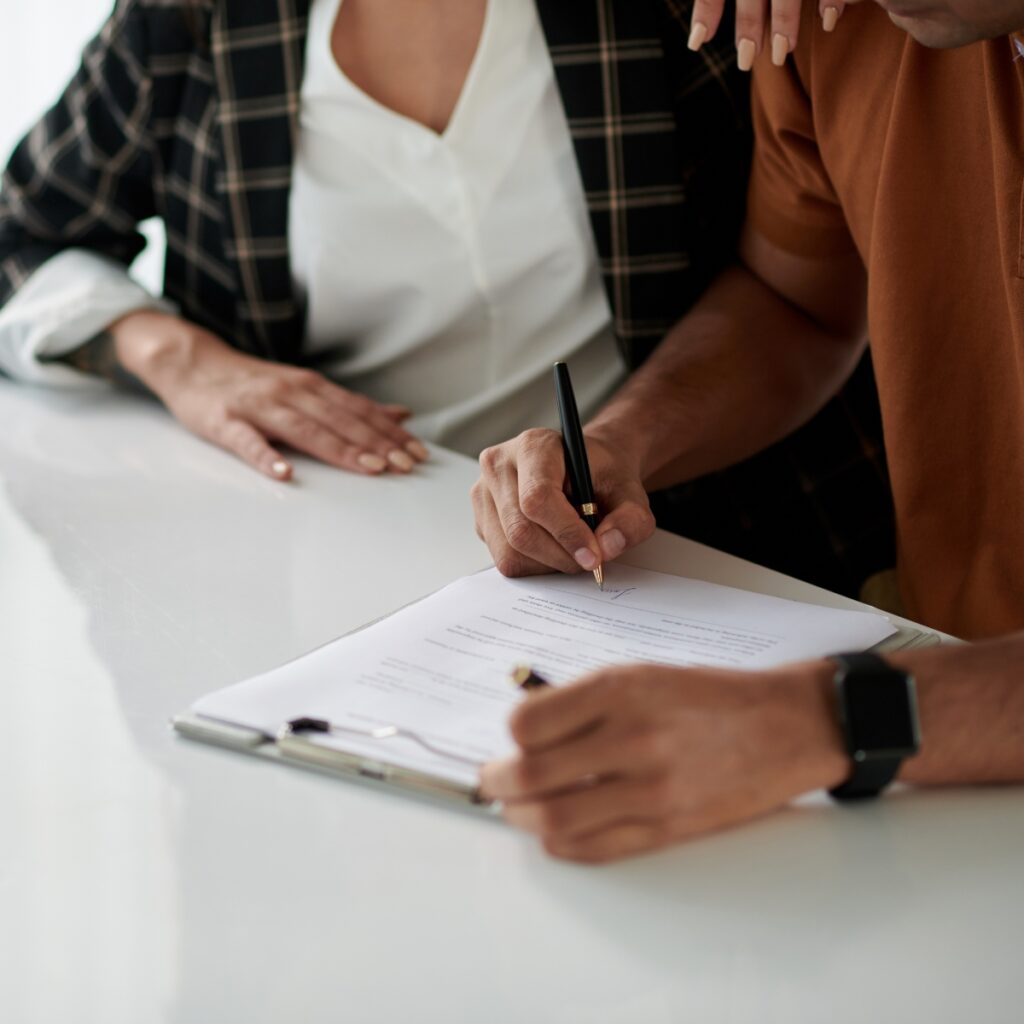 A close-up of a man signing a document on a clipboard with a black and gold pen, while a woman in a plaid blazer sits beside him with her hands resting on the table.